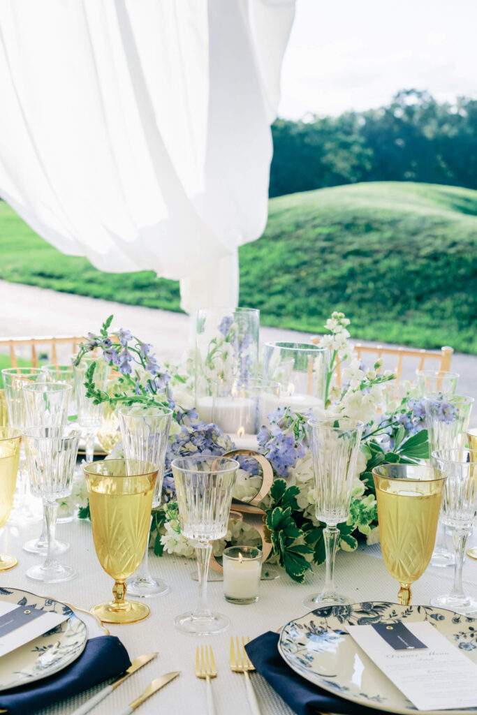 Candlelit tablescape with crystal glassware, navy napkins, and a floral garland centerpiece accented by gold foil table numbers.