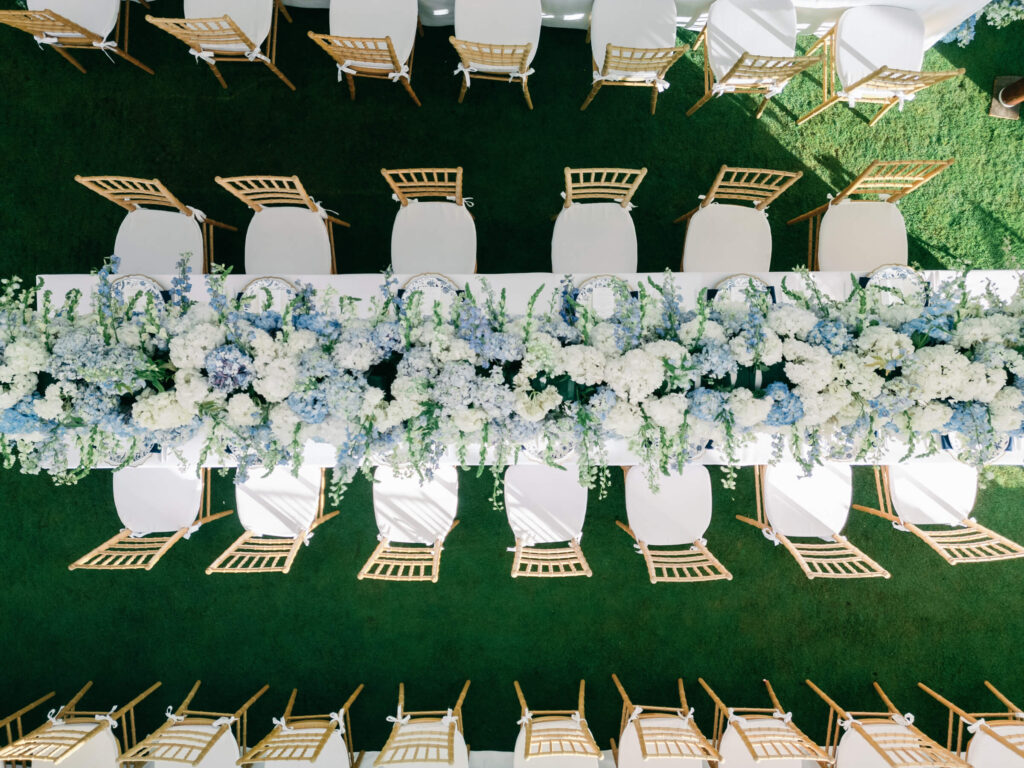 Overhead view of a rectangular reception table covered in lush blue and white flowers, with wooden Chiavari chairs symmetrically aligned on each side.