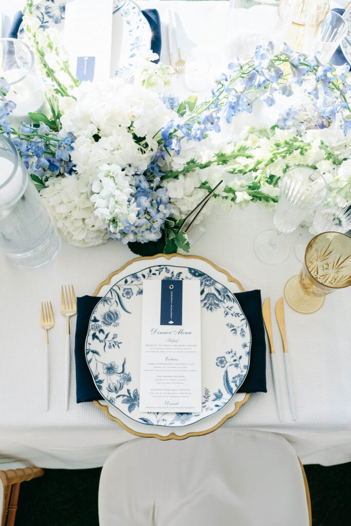 Top-down view of a place setting with navy napkin, gold-rimmed china, blue floral menu card, and surrounding floral centerpiece in ivory and dusty blue.