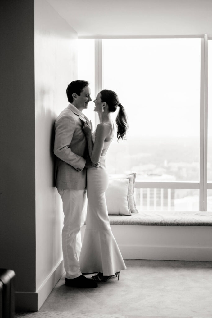 Black and white portrait of a couple standing close near a window, smiling at each other, the bride’s hand gently on the groom’s chest.