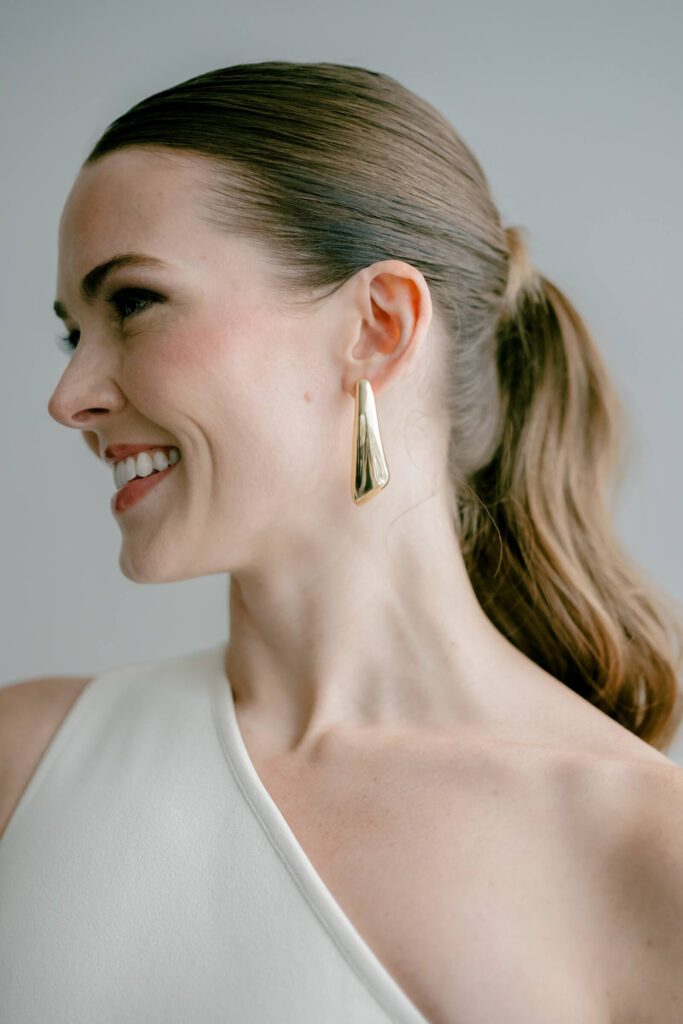 Close-up of the bride smiling softly, showing her sleek ponytail and sculptural gold earring with a clean background.