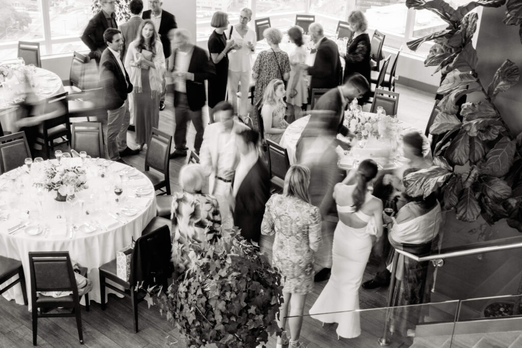 Overhead black and white view of rehearsal dinner guests mingling around round tables with elegant floral centerpieces.
