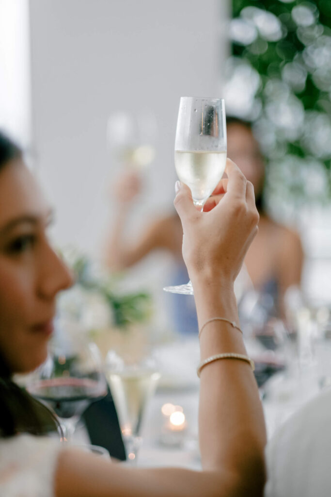 Guest’s hand raising a champagne glass for a toast, with candlelight and floral arrangements softly blurred in the background.