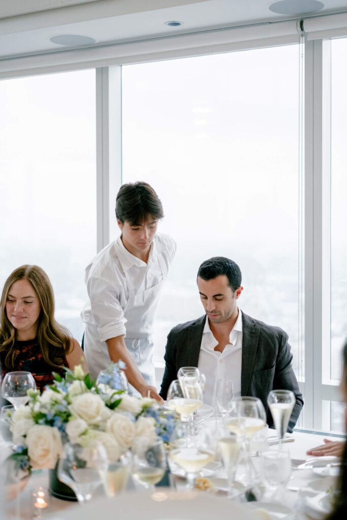 Server placing a plate in front of a guest at a beautifully set reception table adorned with white and blue florals.