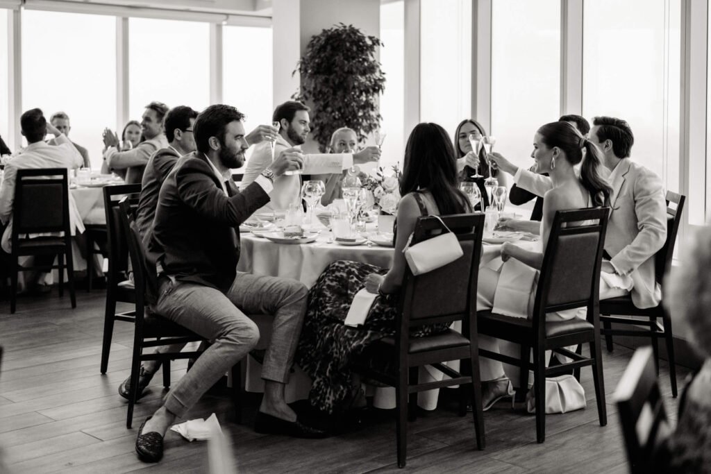Black and white image of guests seated at a round table, raising glasses for a toast, with the couple among them.