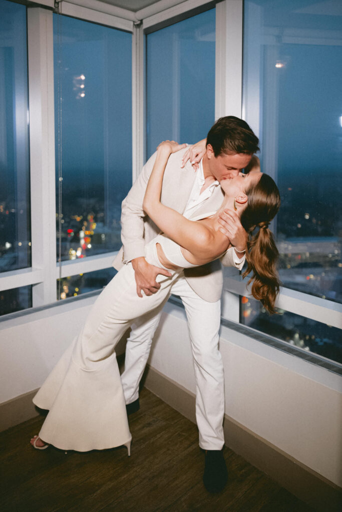 The couple shares a dramatic kiss in a corner window at night, the city lights glowing outside behind them.
